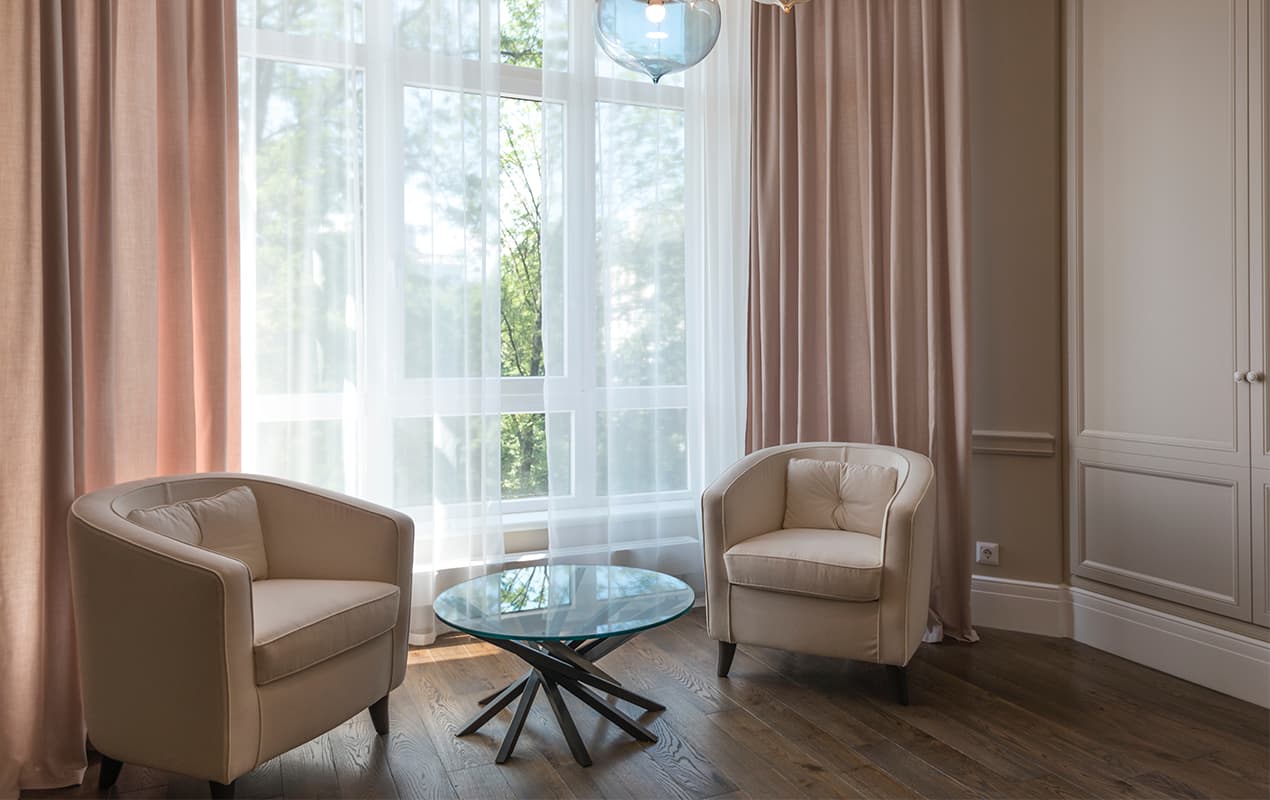 Baby pink floor to ceiling curtains, beige armchairs in front of windows
